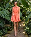 Exquisite Bows Women's Dress in coral styled in a tropical garden, featuring tonal bows and a structured pleated skirt.